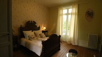 A bedroom with a wooden bed, a bedside table with a lamp, a glass table, and a radiator.