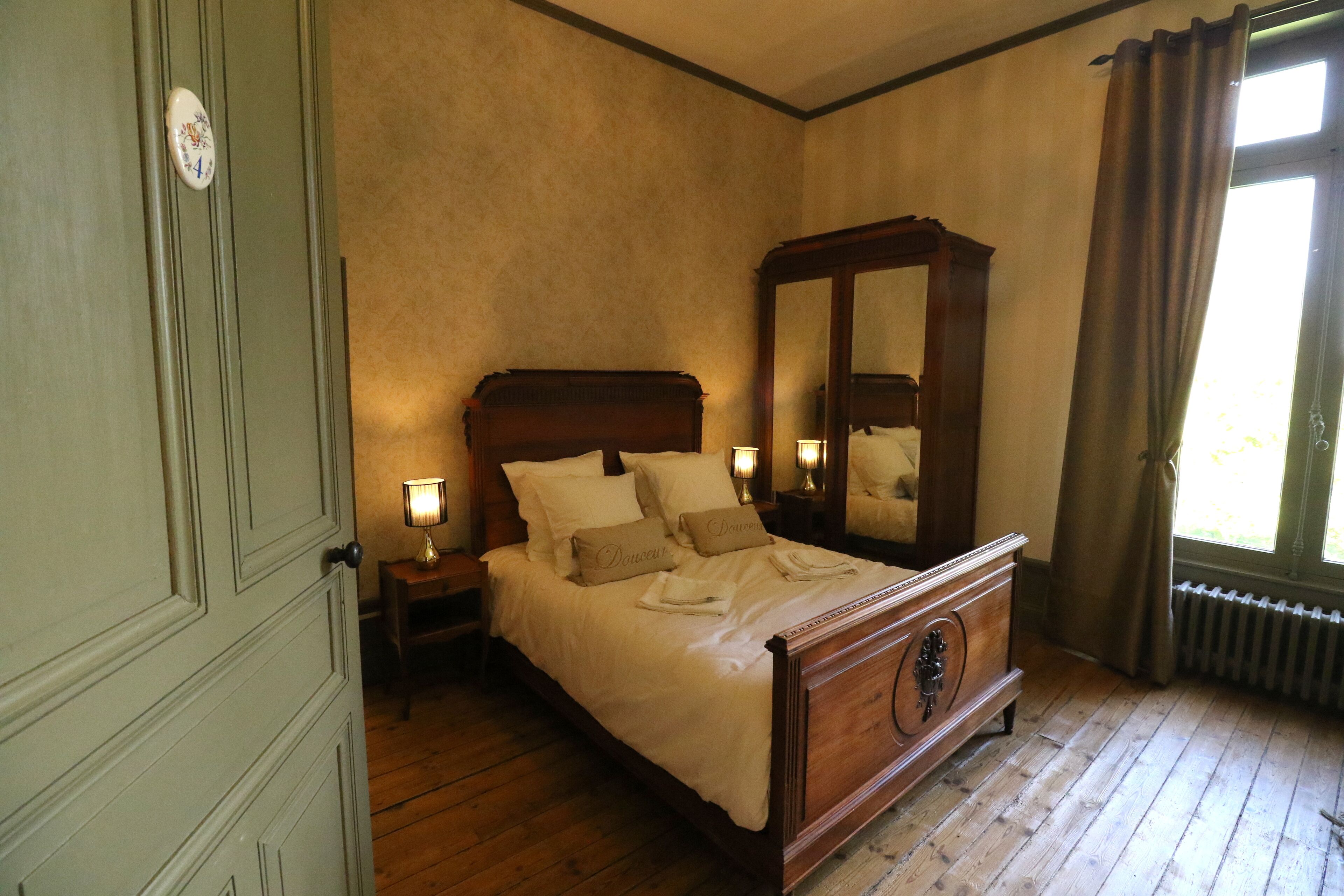 A bedroom with a wooden bed, a nightstand with a lamp, a large mirror, and a window with curtains.