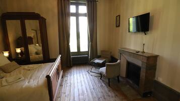 Un dormitorio con piso de madera, una cama con cabecera, una silla, chimenea, televisor y ventana con cortinas.