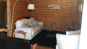 Een knus houten blokhut interieur met een witte leren bank, twee gestreepte kussens, een glazen salontafel en een houten eettafel met stoelen.