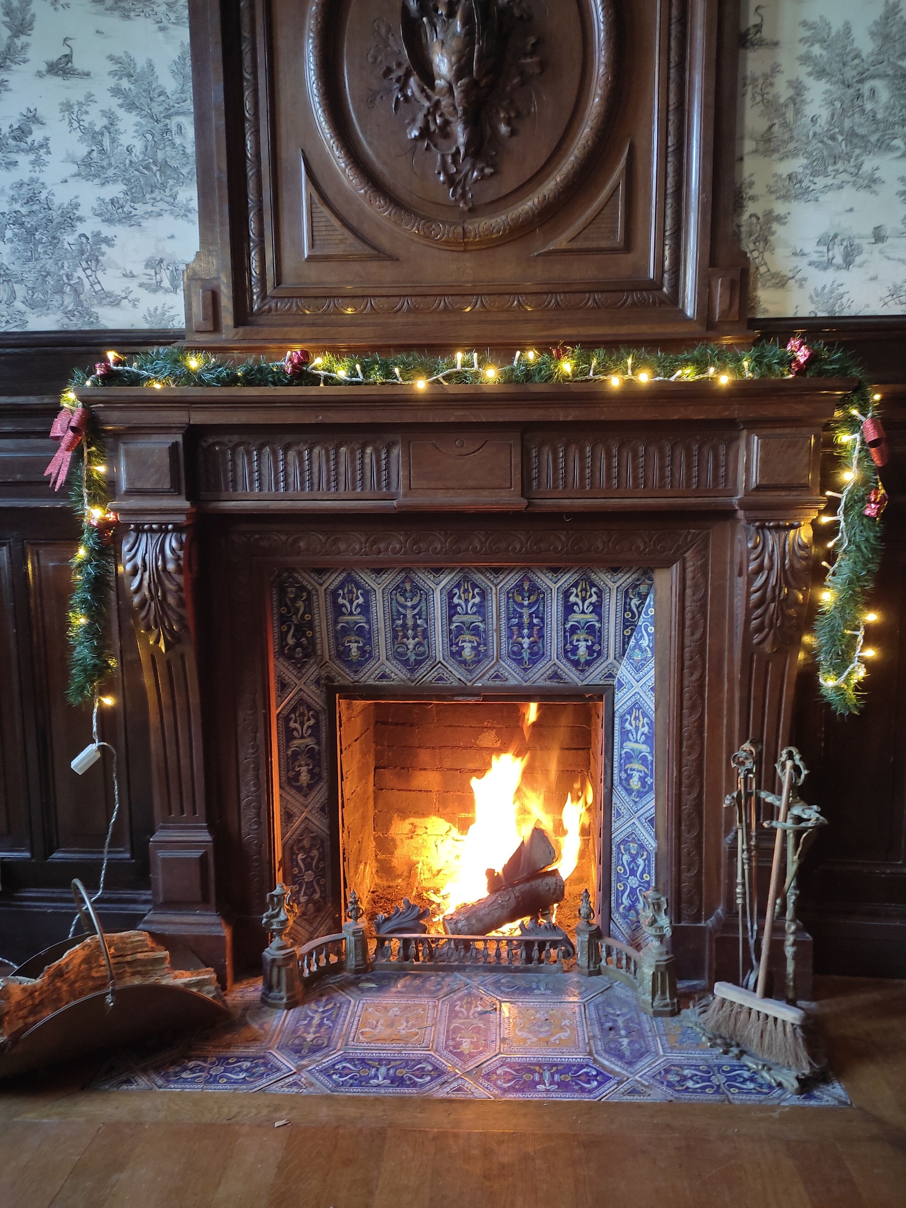 A fireplace with a fire, surrounded by ornate wooden mantel and a patterned tile floor.