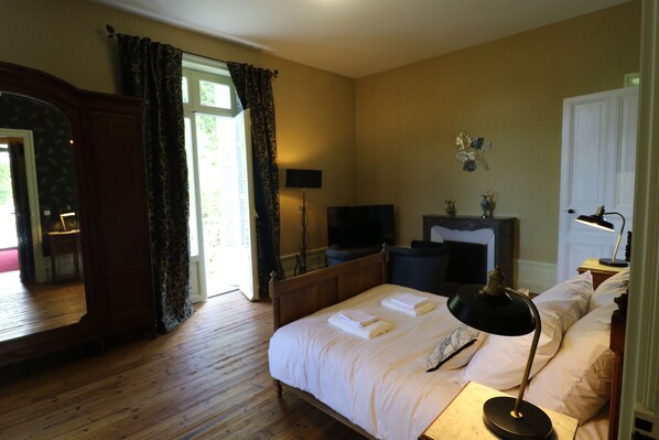 A bedroom with a wooden floor, a bed with white linens, a bedside table with a lamp, a television, and a fireplace.