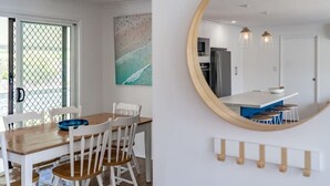 A dining area with a wooden table and chairs, a kitchen in the background, and a large mirror on the wall.