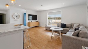 A modern living room with a sofa, a small round table, a TV, and a kitchen area with a counter and stools.
