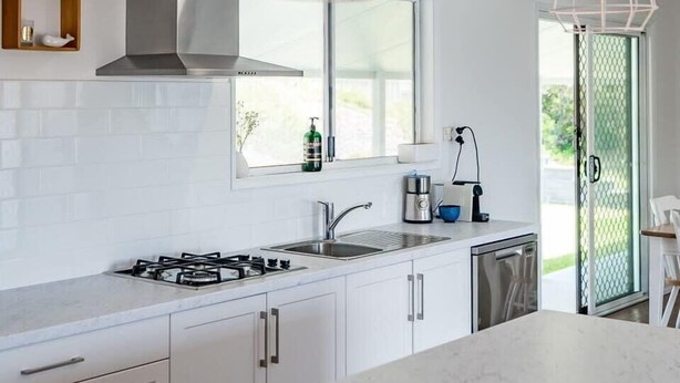 A modern kitchen with white cabinetry, a gas stove, a stainless steel range hood, pendant lighting, and a window providing natural light.