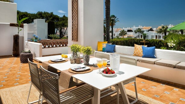 Een terras met een eettafel, stoelen en een bank met kussens. Op de tafel staan planten, een vaas en een schaal met fruit.