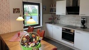 A modern kitchen with white cabinets, a dining table with chairs, a hanging light fixture, and a window with a view of a house and greenery.