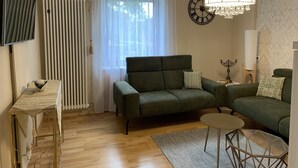 A living room with a green sofa, a small round table, a TV on a stand, a radiator, a window with sheer curtains, a wall clock, and a chandelier.