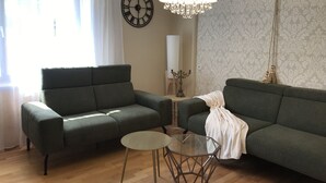 A living room with a green sofa, a glass coffee table, a chandelier, and a wall clock.
