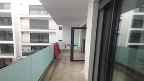 Ein schmaler Korridor mit gefliestem Boden, einer Glastür auf der rechten Seite und Blick auf ein Gebäude mit mehreren Balkonen.