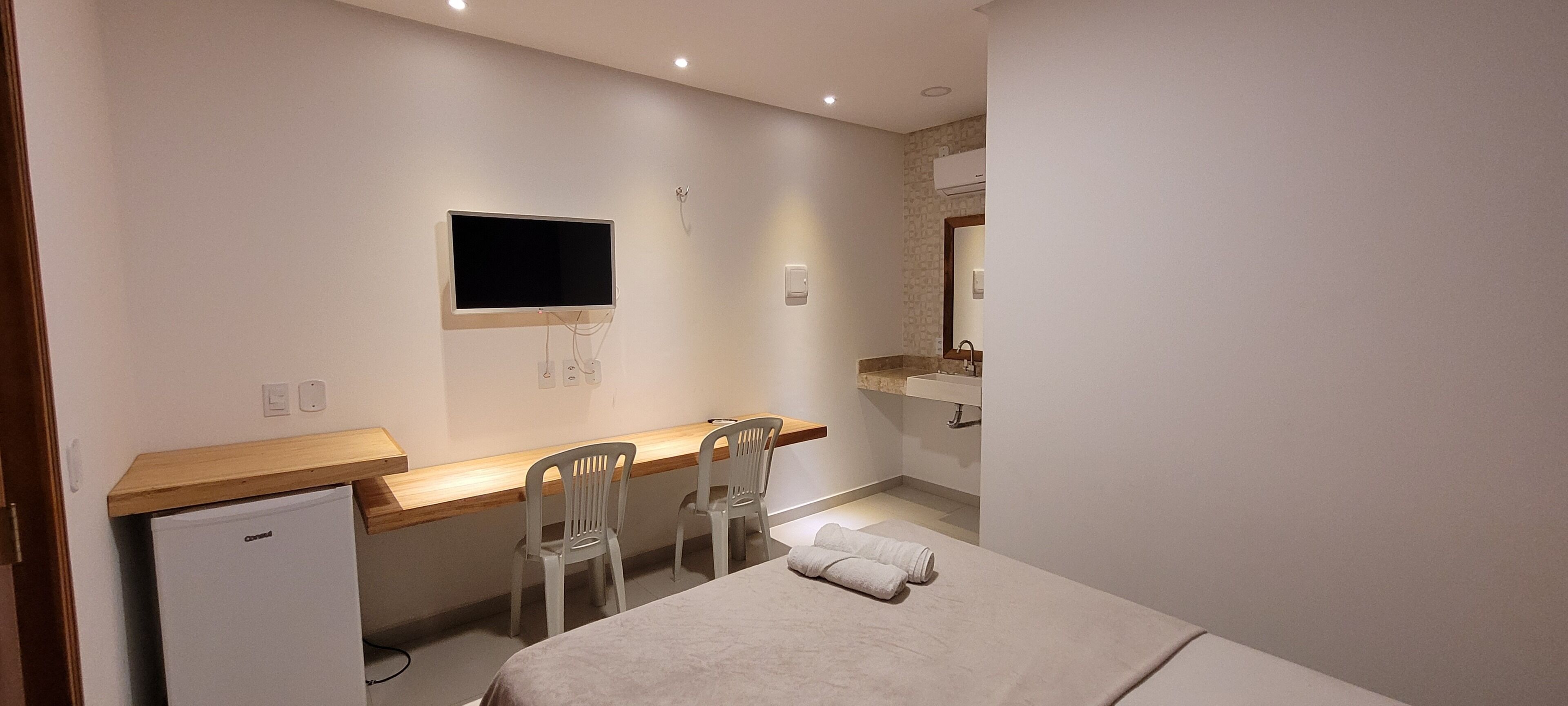A hotel room with a bed, a desk with two chairs, a television mounted on the wall, a small refrigerator, and a bathroom with a sink and mirror.