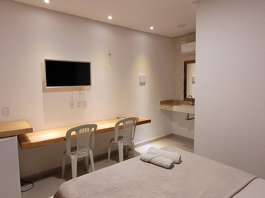 A hotel room with a bed, a desk with two chairs, a television mounted on the wall, a small refrigerator, and a bathroom with a sink and mirror.