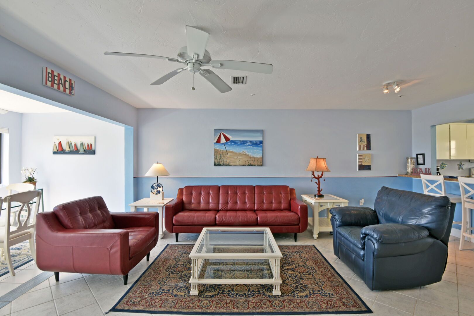 A living room with a red leather sofa, a blue leather armchair, a glass coffee table, and a ceiling fan. There is a painting of a beach scene on the wall, a lamp on a side table, and a small kitchen area in the background.