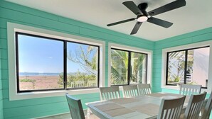 Un coin repas avec une table et des chaises, de grandes fenêtres donnant sur la plage et un ventilateur de plafond.