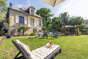 A spacious outdoor area with a lounge chair, a dining table with a bottle and a plate, and a pergola with chairs.
