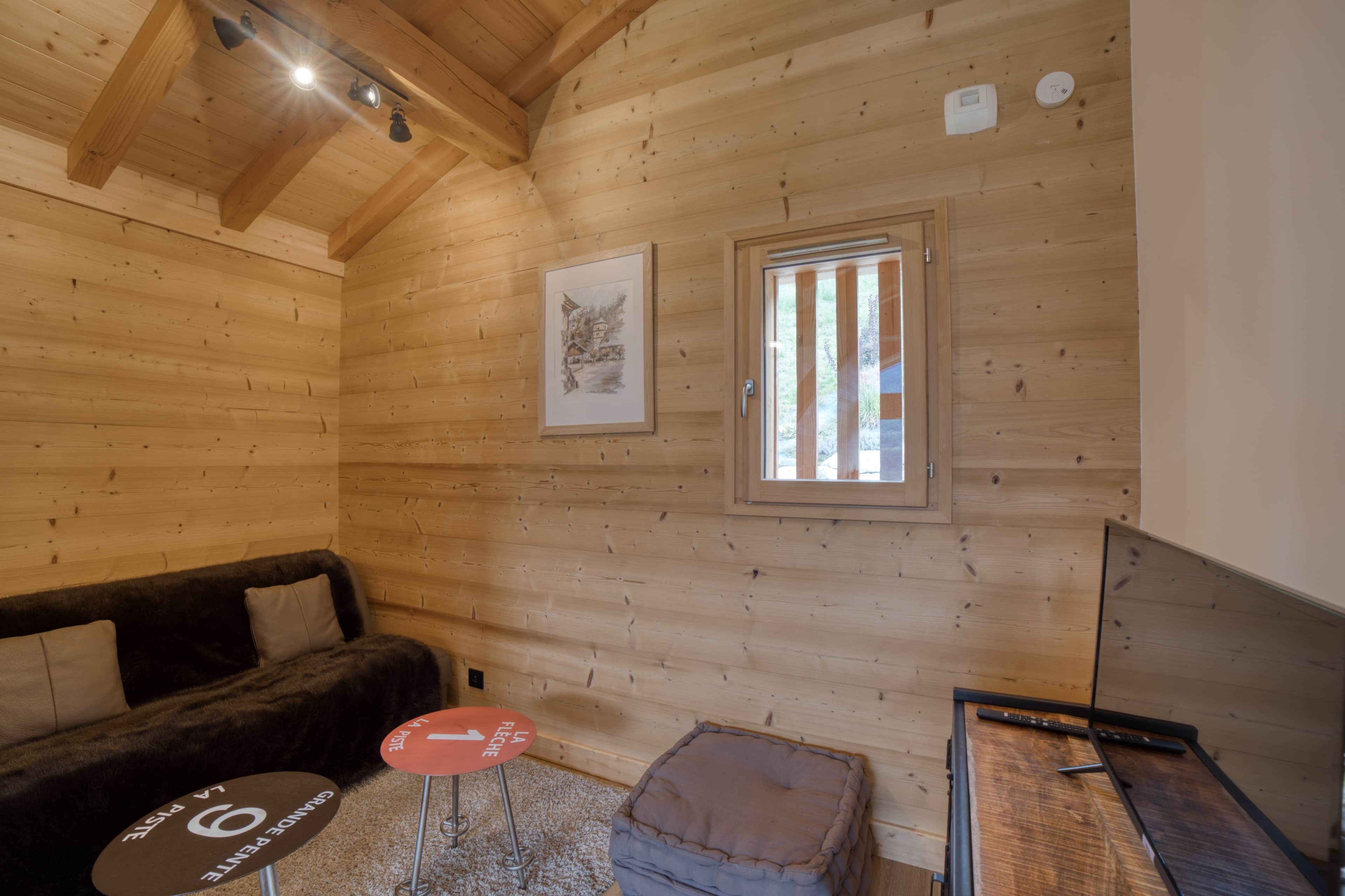 A cozy cabin interior with a wooden ceiling, a framed picture on the wall, a window with vertical blinds, a sofa, a small round table, and a television.