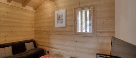 Ein gemütliches Blockhaus-Interieur mit Holzbalkendecke, einem gerahmten Bild an der Wand, einem Fenster mit vertikalen Rollläden, einer Couch, einem kleinen runden Tisch und einem Fernseher.