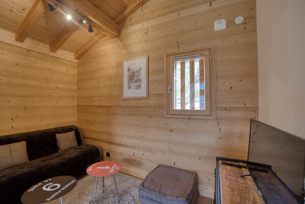 L’intérieur d’un chalet accueillant, avec un plafond en bois, un tableau encadré au mur, une fenêtre avec des stores verticaux, un canapé, une petite table ronde et un téléviseur.