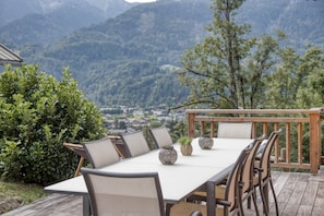 An outdoor dining area with a table and chairs, surrounded by a wooden deck and a view of a mountainous landscape.