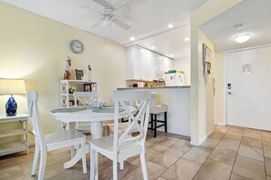 Uma área de refeições com cadeiras brancas e uma mesa redonda, uma cozinha com armários brancos e um ventilador de teto.