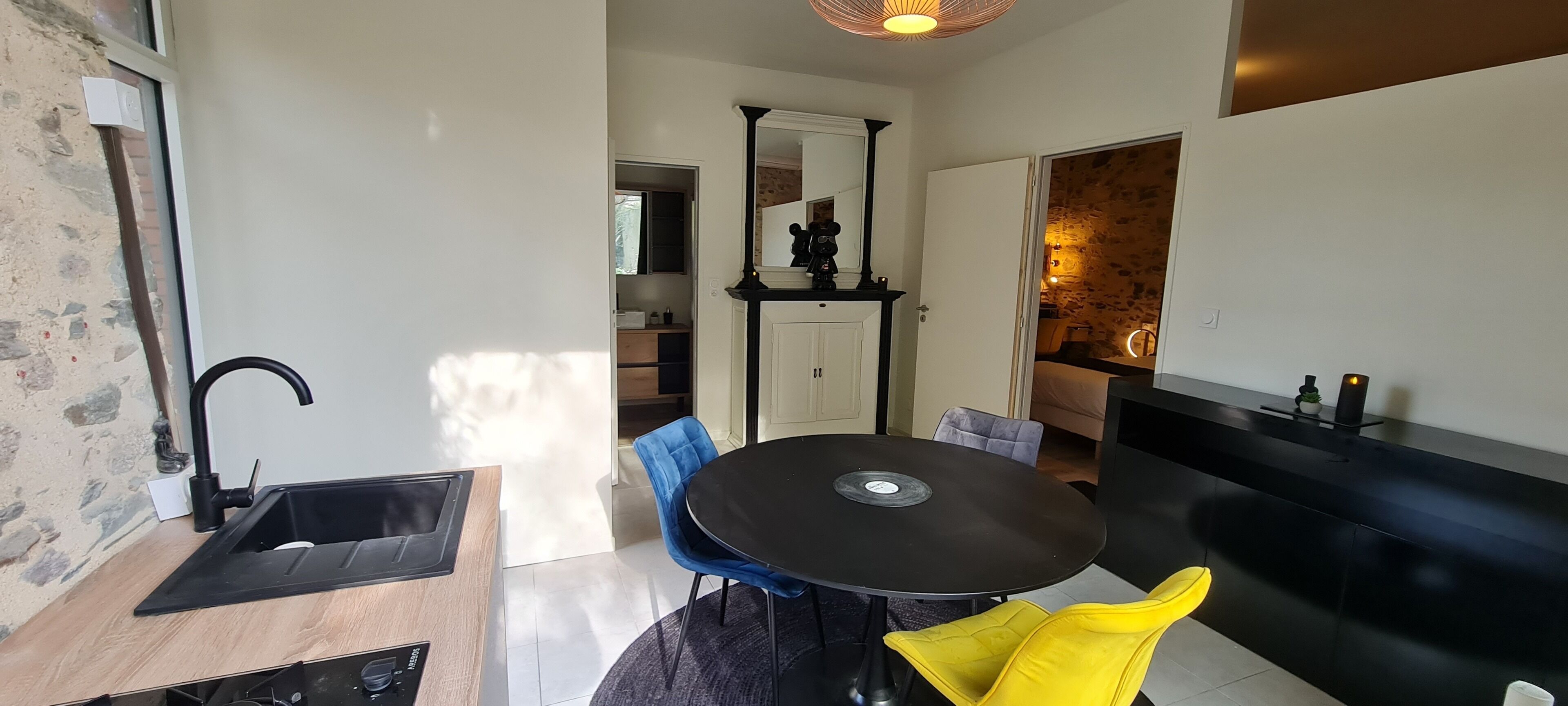 Et moderne køkken med en sort vask, en træbordplade og et indbygget komfur. Der er et spiseområde med et rundt bord og to stole, en blå og en gul. Et sort skab med et spejl er synligt i baggrunden.