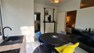 Et moderne køkken med en sort vask, en træbordplade og et indbygget komfur. Der er et spiseområde med et rundt bord og to stole, en blå og en gul. Et sort skab med et spejl er synligt i baggrunden.