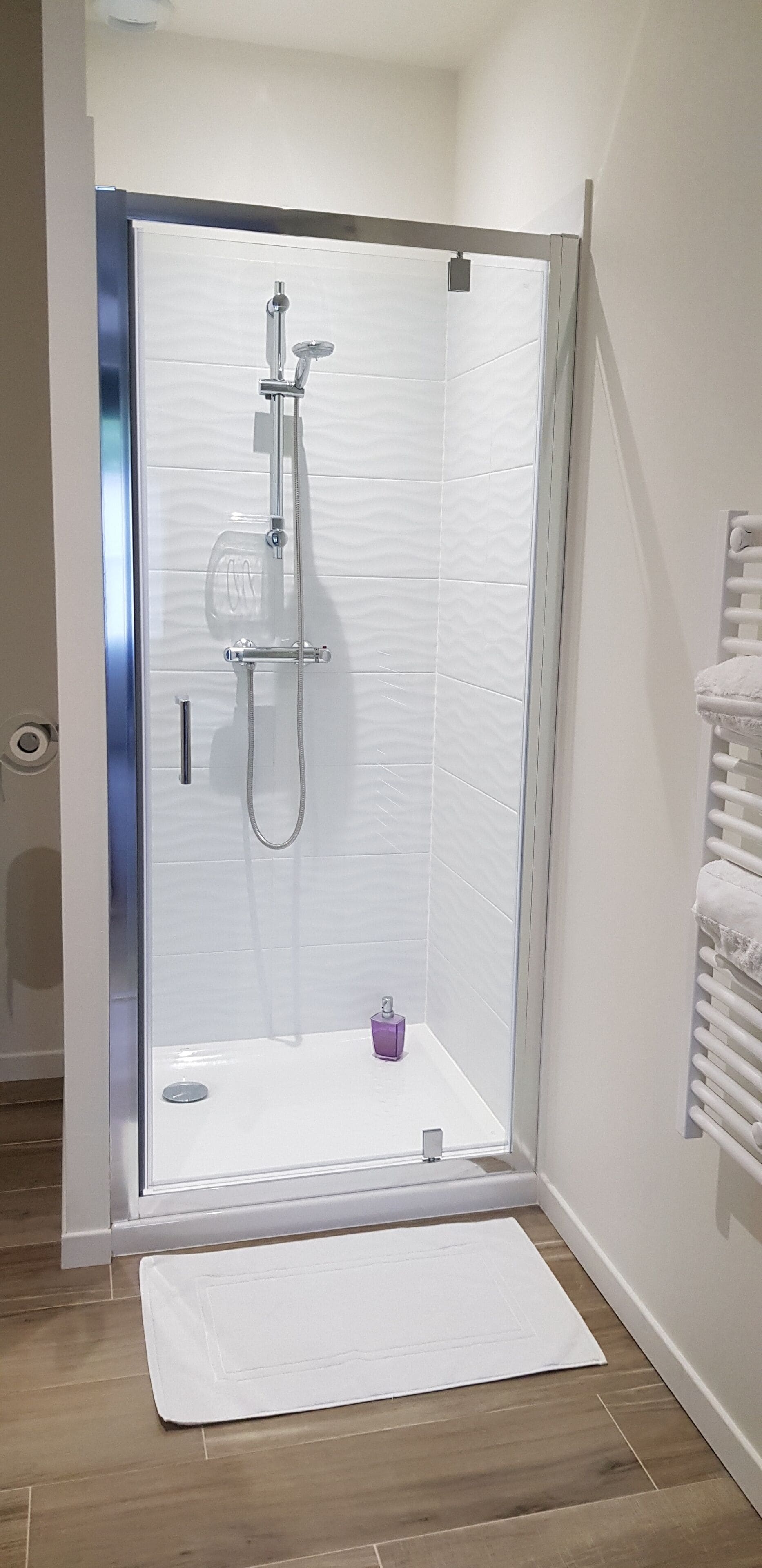 Una ducha con cerramiento de vidrio, pared de azulejos blancos, toallero blanco y un tapete en el suelo.