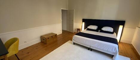 Un dormitorio con cama, alfombra blanca, suelo de madera, una silla amarilla y una lámpara colgante.