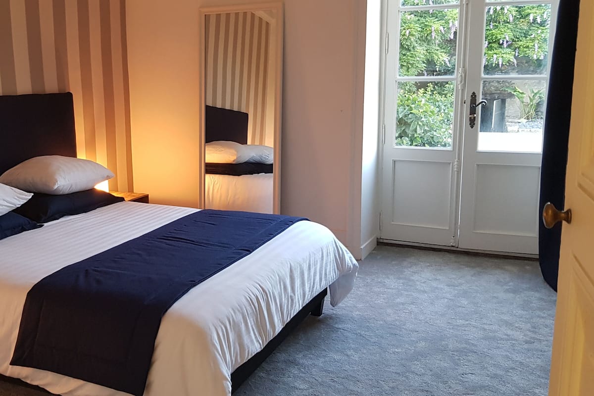 Una cama bien hecha con ropa de cama blanca y azul marino, una mesita de noche con una lámpara y vistas a la vegetación a través de una puerta.