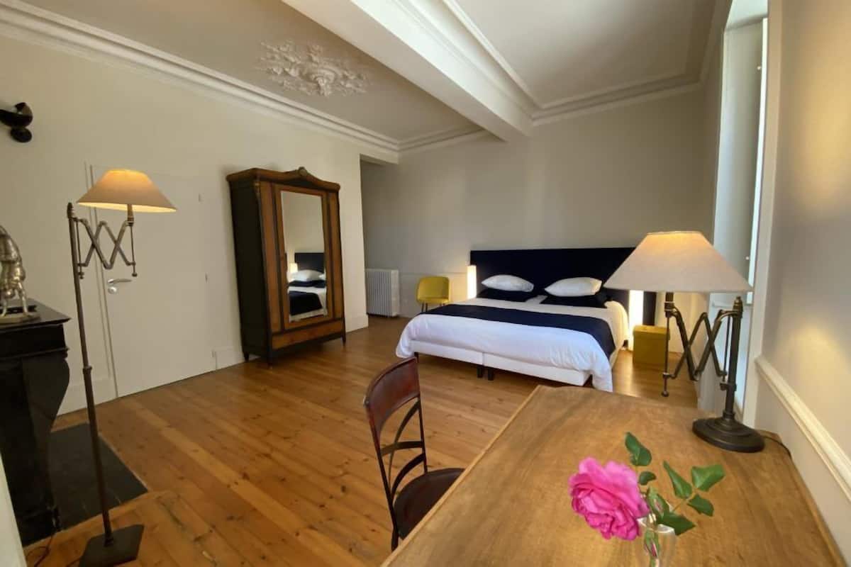 Un dormitorio con cama, lámparas de noche, una mesa de madera, una silla, un espejo y un florero con una flor.