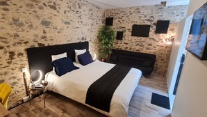 Een slaapkamer met een stenen muur, een bed met een zwart hoofdeinde, een nachtkastje, een stoel, een plant en een bank.