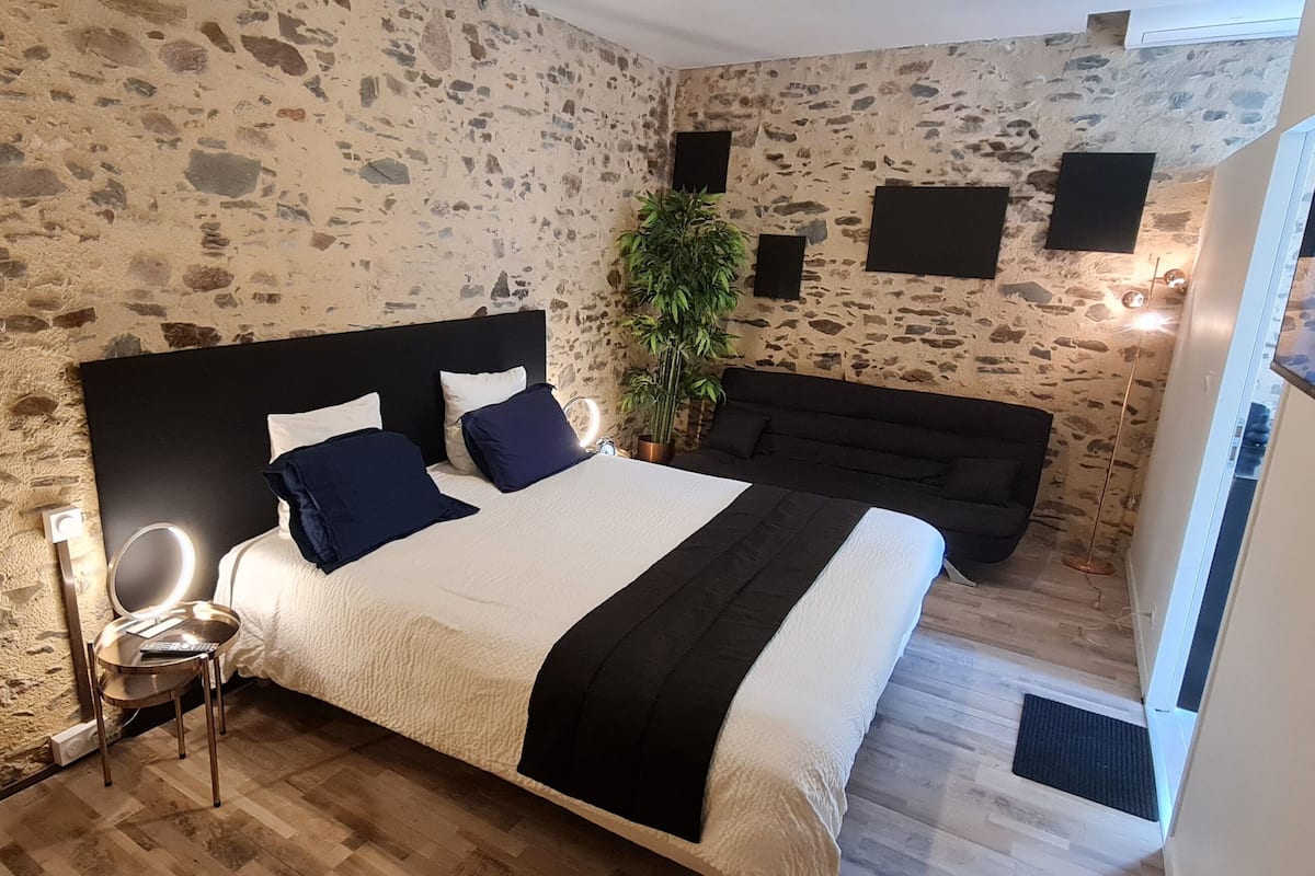 Un dormitorio con pared de piedra, cama con cabecera negra, mesita de noche, una silla, planta y sofá.