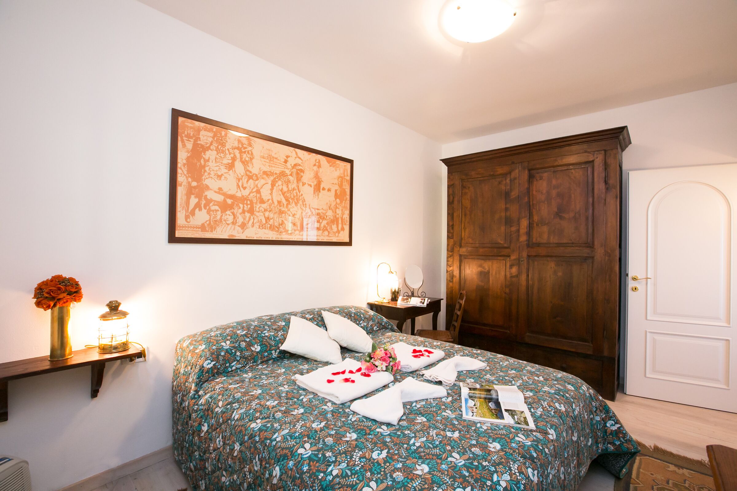 A neatly made bed with floral bedding, a bedside table with a lamp, and a large framed picture on the wall.