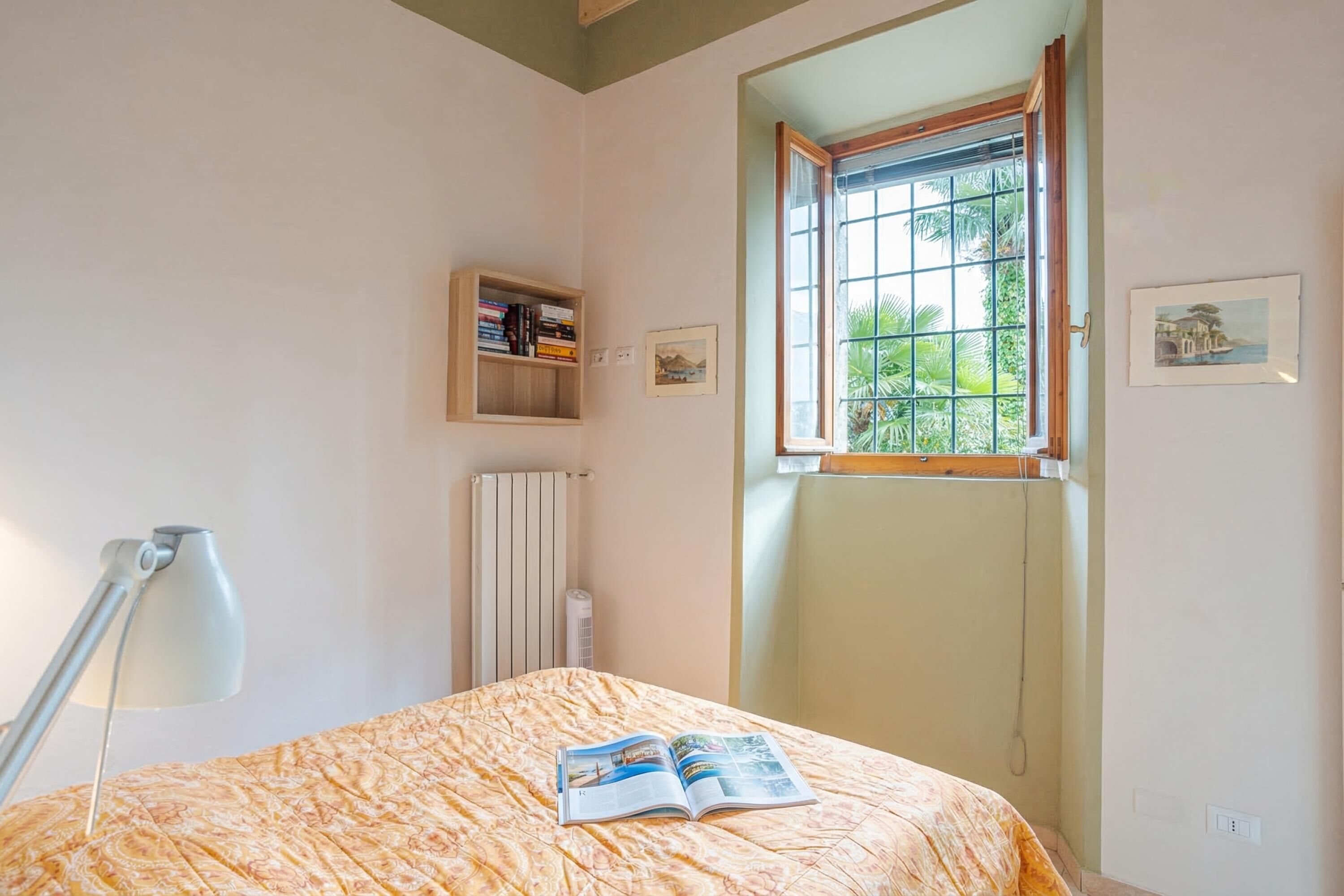 A bedroom with a bed, a window, a radiator, a shelf with books, and a wall-mounted picture.