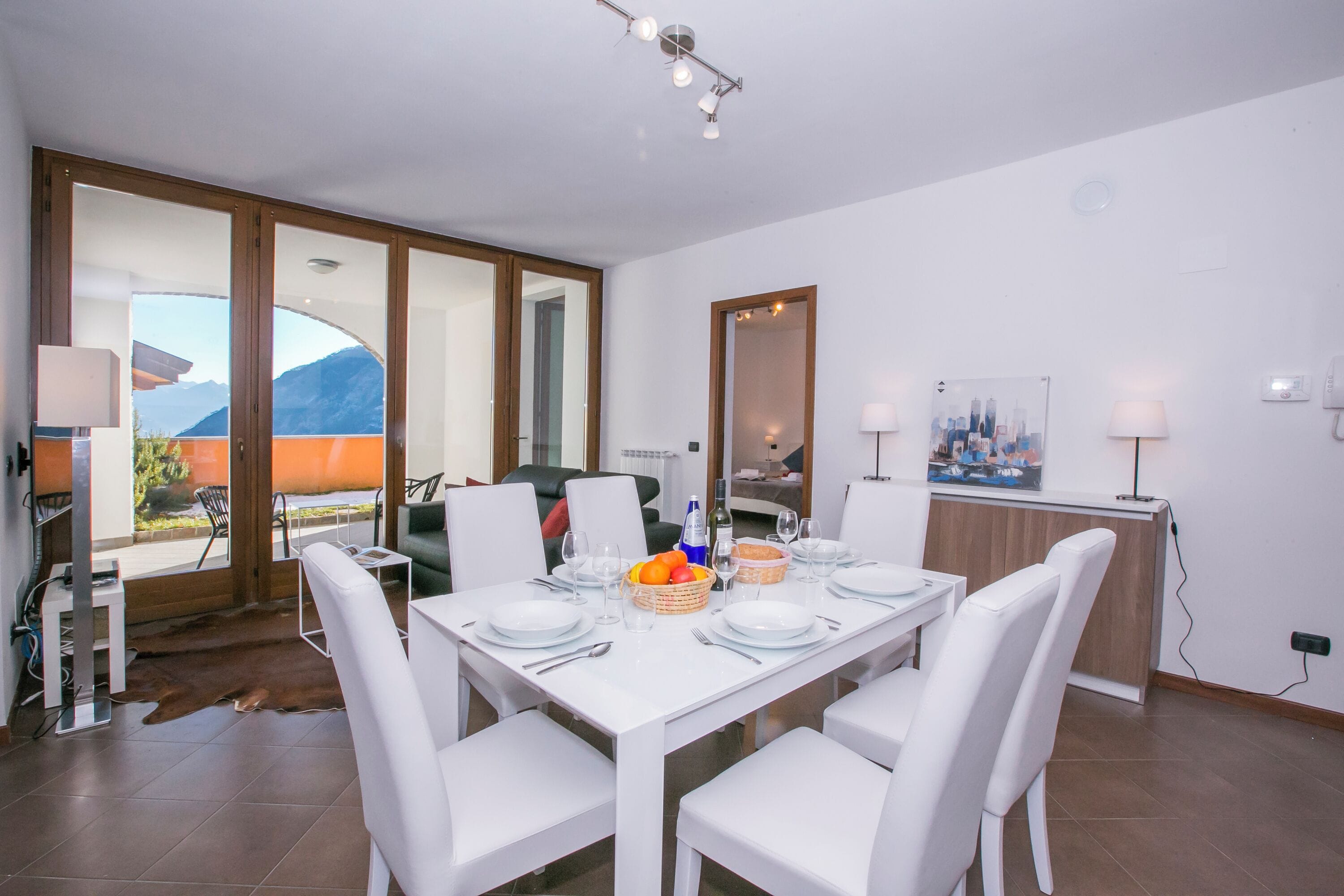 A dining area with a white table set for four, chairs, a fruit basket, and a cityscape painting on the wall.