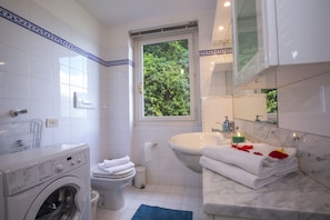 A modern bathroom with a washing machine, toilet, sink, and a window with a view of greenery.