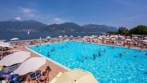 A large outdoor swimming pool with people swimming and sunbathing, surrounded by umbrellas and beach chairs, with a view of mountains and the sea in the background.