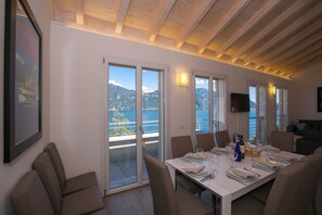 A dining area with a table set for four, large windows offering a view of mountains and a body of water, and a ceiling with exposed wooden beams. - Paradiso di Menaggio Penthouse (Menaggio)