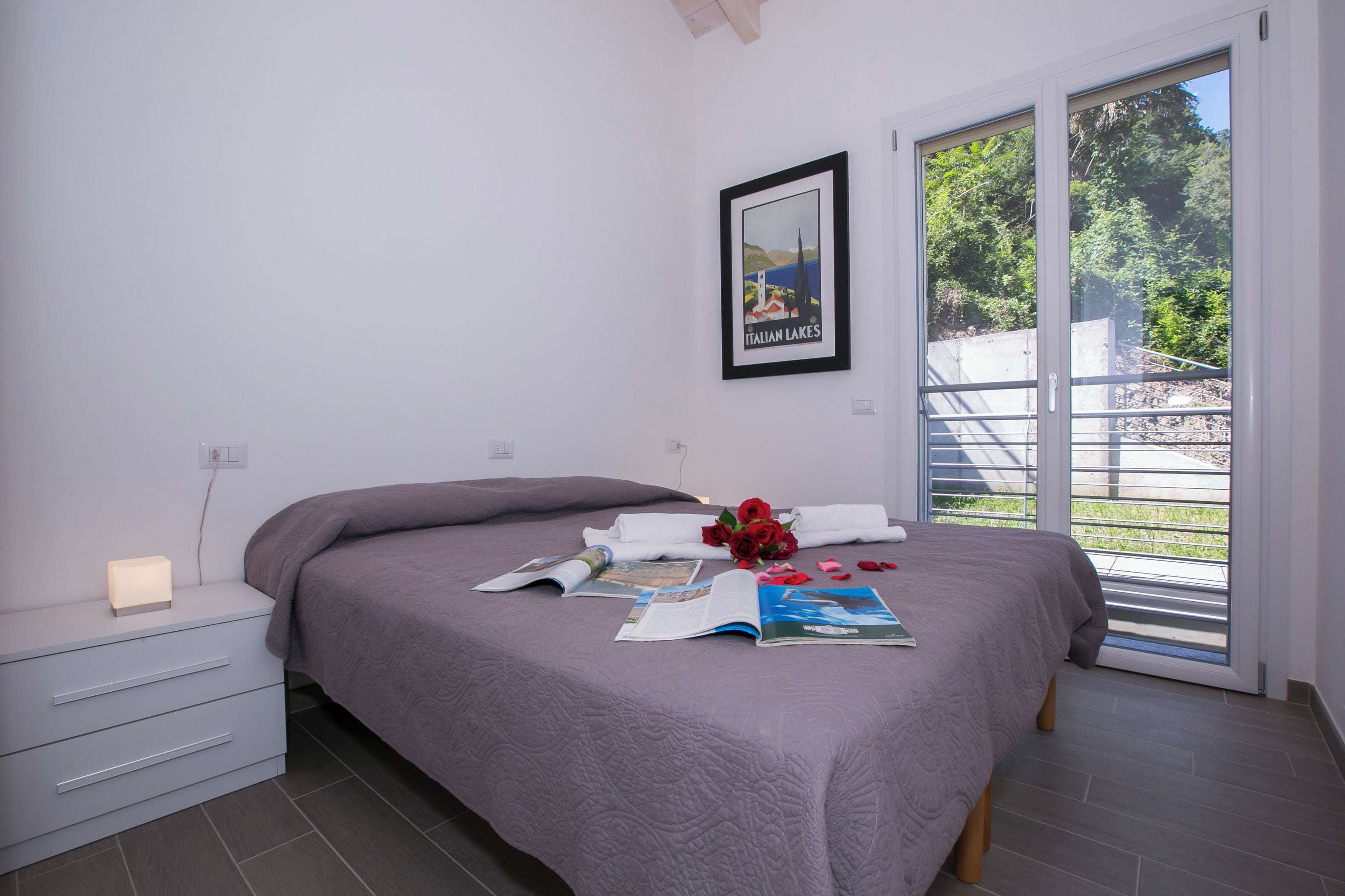 A bedroom with a bed, a bedside table with a lamp, a framed picture on the wall, and a window with a view of greenery.