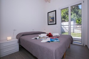 A bedroom with a bed, a bedside table with a lamp, a framed picture on the wall, and a window with a view of greenery. - Paradiso di Menaggio Penthouse (Menaggio)