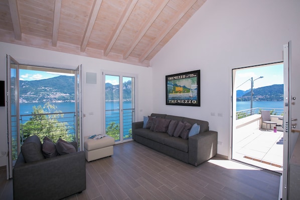 A spacious living room with a grey sofa, a small ottoman, and a large window offering a view of a mountainous landscape. - Paradiso di Menaggio Penthouse (Menaggio)