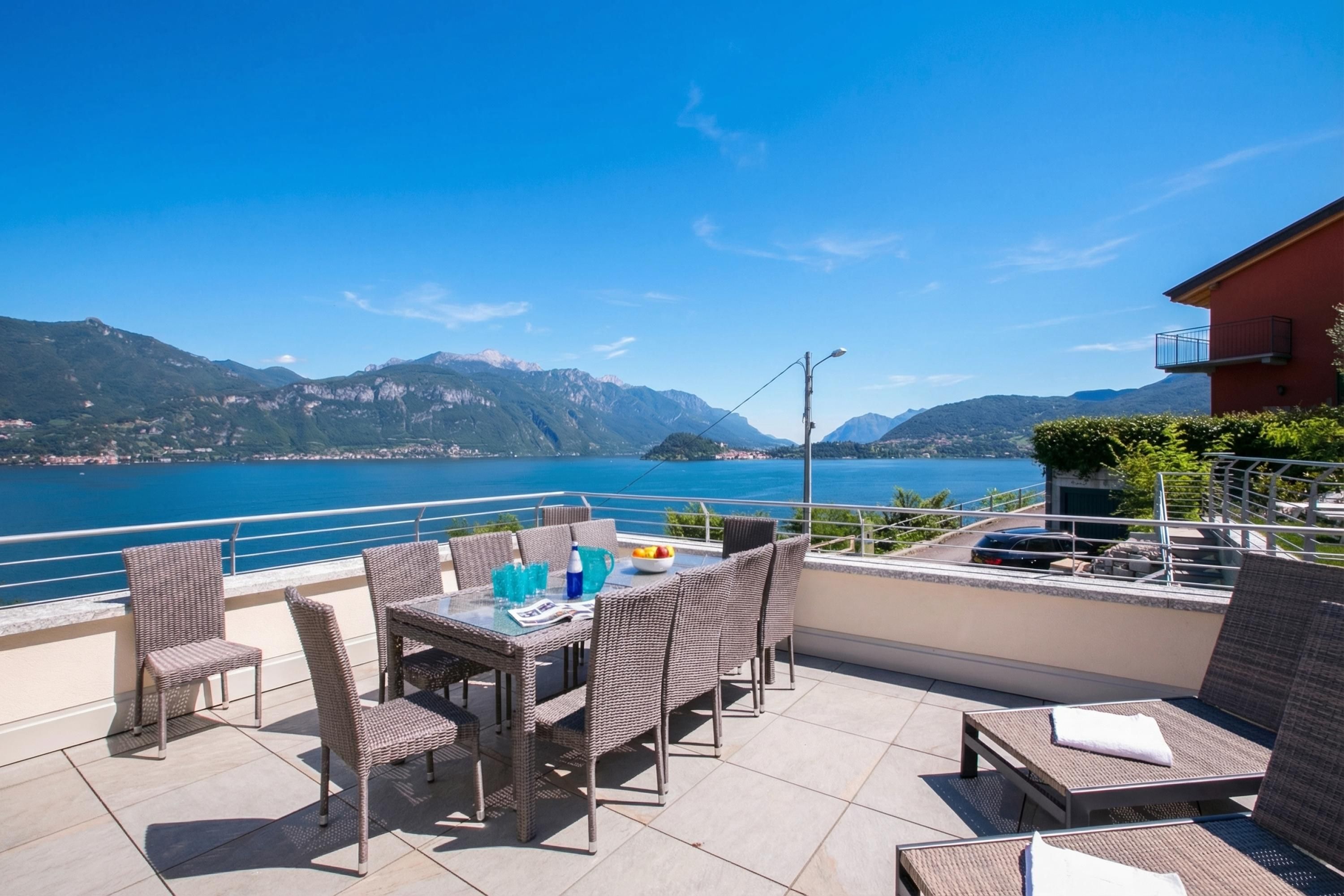 Une terrasse meublée de mobilier en osier, une table avec des boissons et une vue sur un lac et des montagnes.