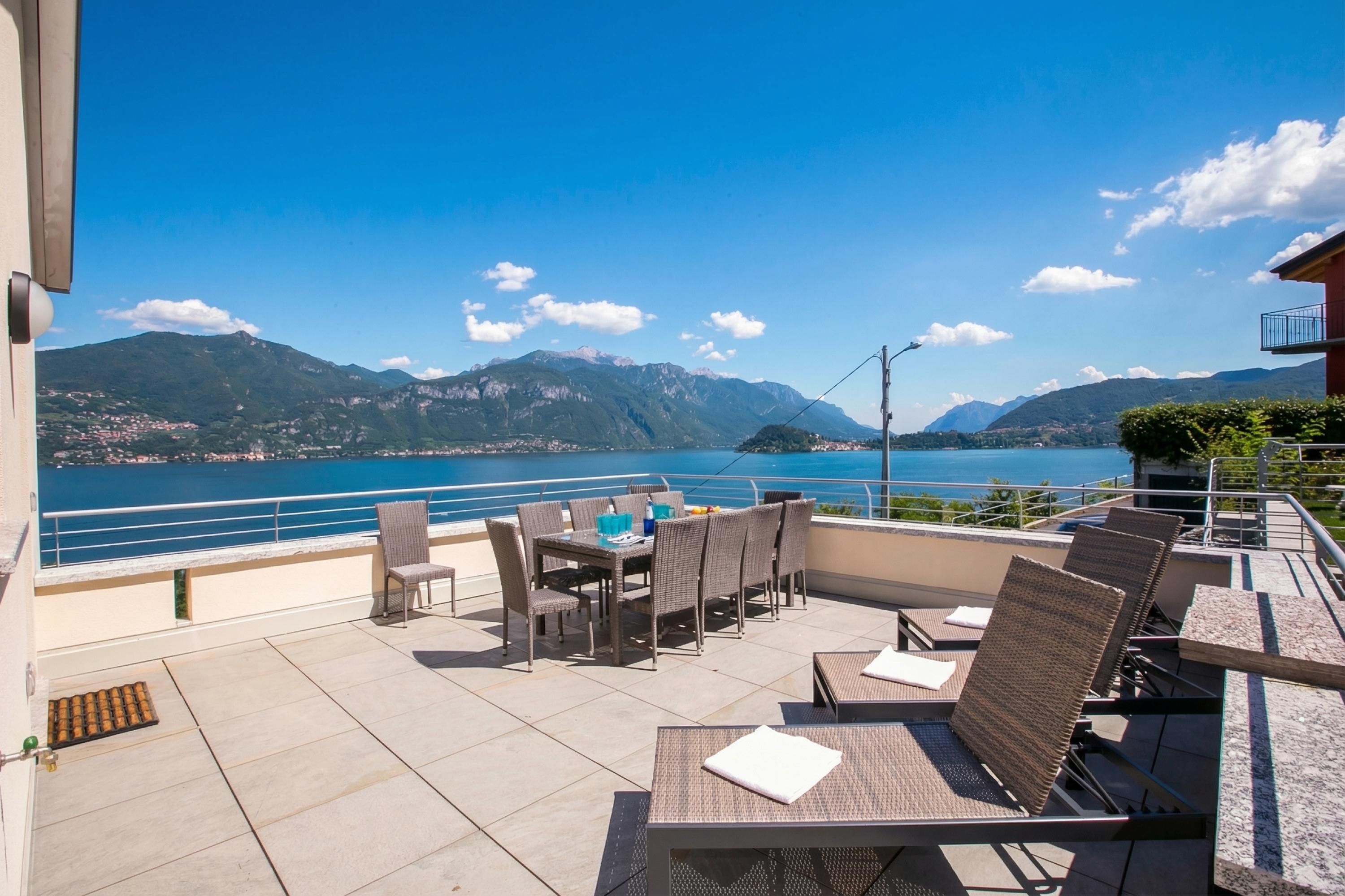 An outdoor terrace with a table and chairs, overlooking a body of water and mountains.
