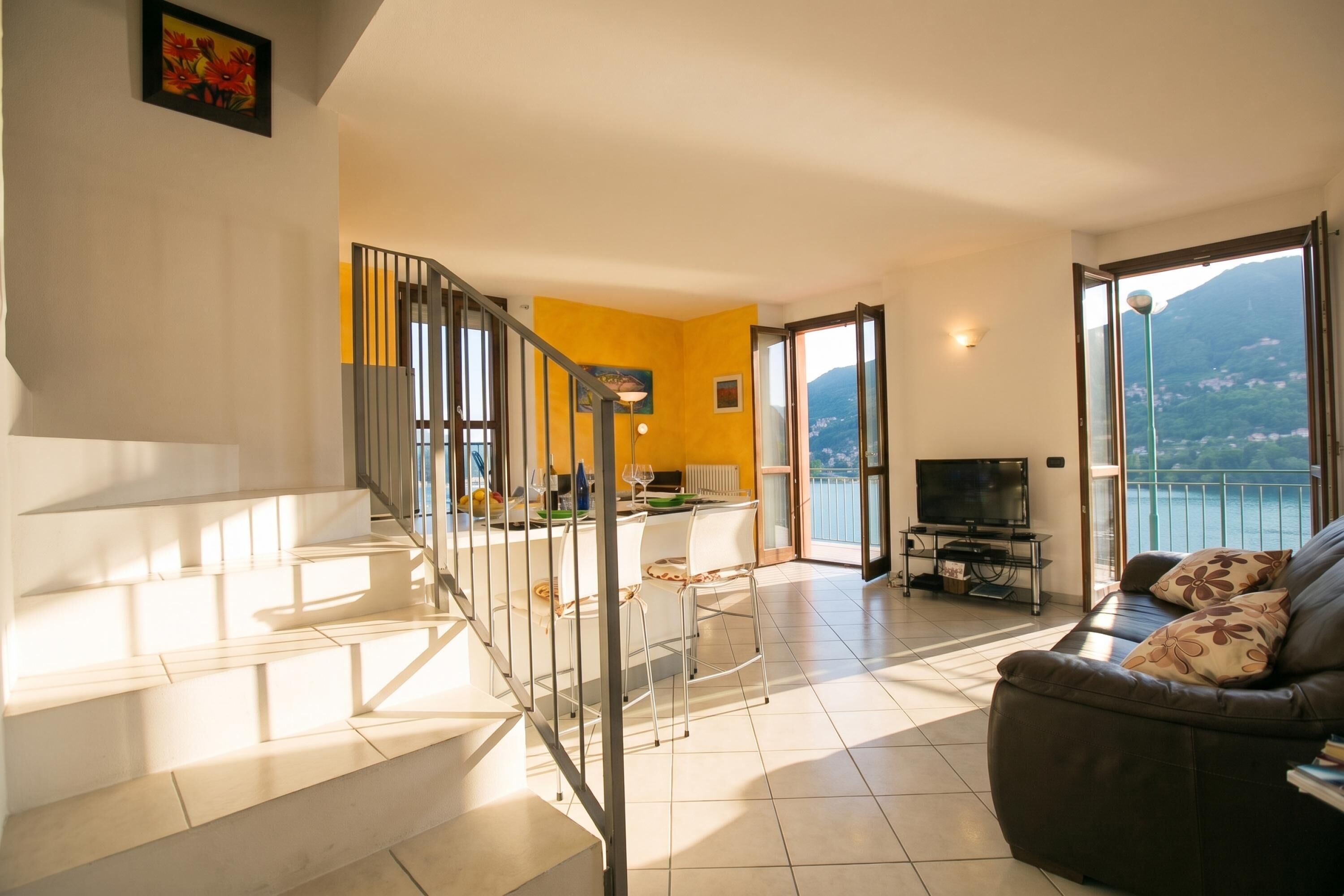 A living room with a sofa, a dining area with a table and chairs, a staircase, and a balcony with a TV.