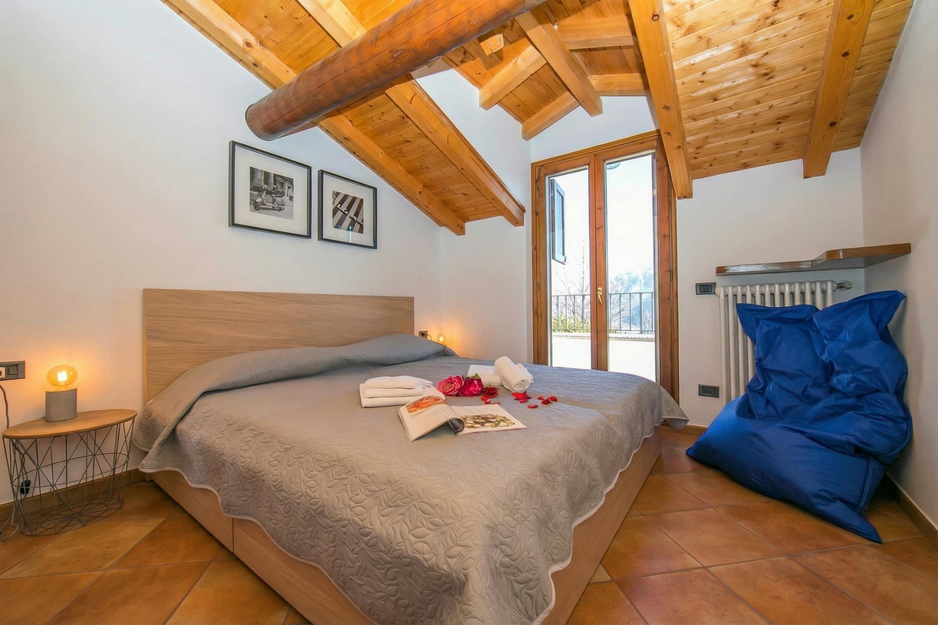 Quarto com teto de madeira, cama com colcha cinza, mesa de cabeceira com abajur e radiador.