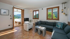 Ein Wohnzimmer mit einer Couch, einem Couchtisch, einem Sessel und einem Fernseher. Eine Tür führt zu einem Außenbereich.