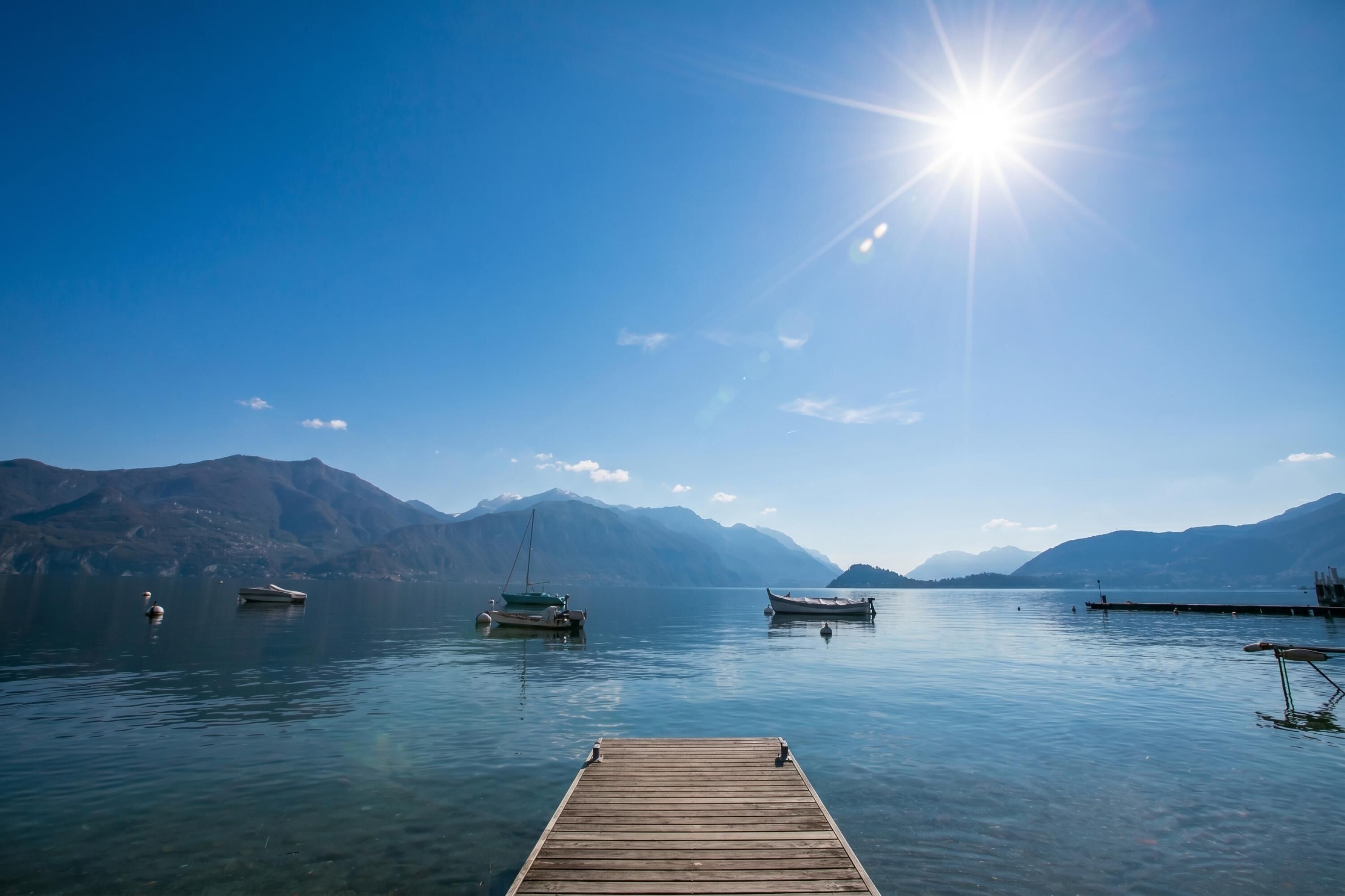 A wooden dock extending into calm water with boats anchored nearby and mountains in the background.