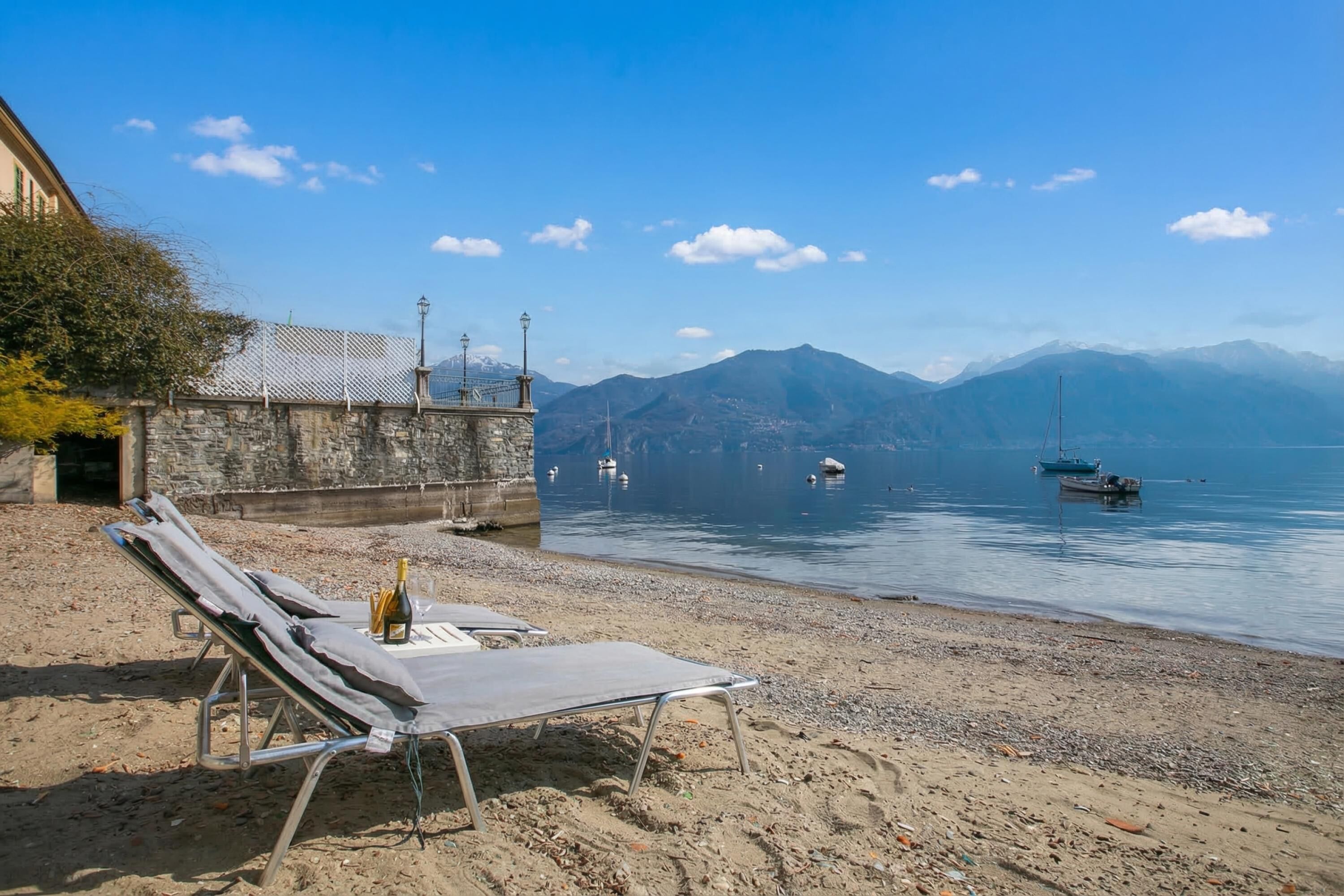 A beach with lounge chairs and a bottle of wine, a stone wall, and boats on calm water with mountains in the background.