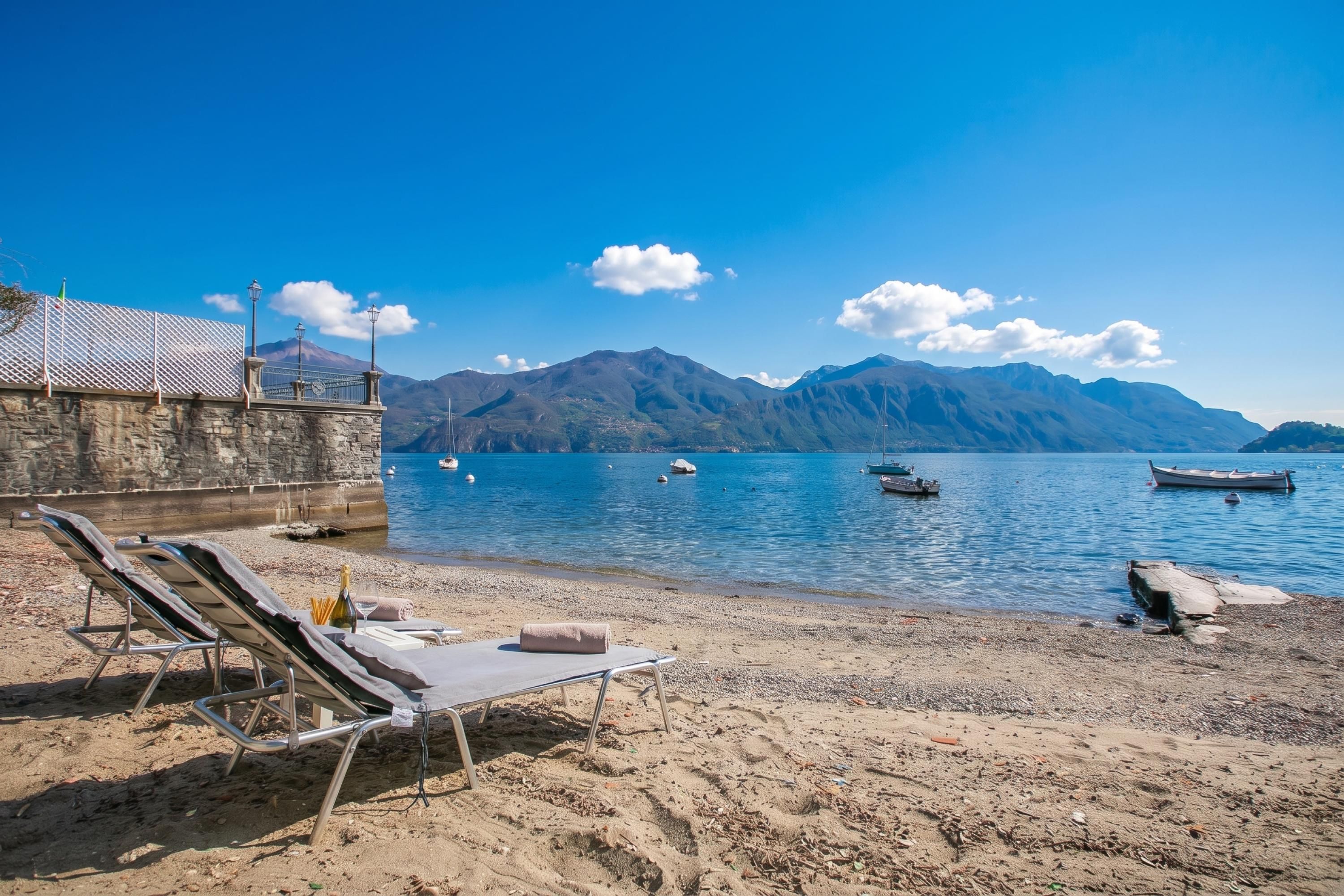 Una playa con sillas de descanso, un muro de piedra y barcos sobre un lago tranquilo, con montañas al fondo.