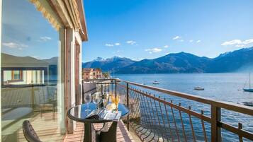 Un balcon avec une table et des chaises, donnant sur un lac où l’on aperçoit des bateaux, avec des montagnes en arrière-plan.